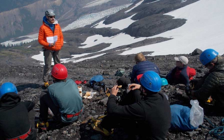 An instructor stands and speaks to students sitting on the ground. They appear to be at high elevation by the snow in the background and the lack of greenery.